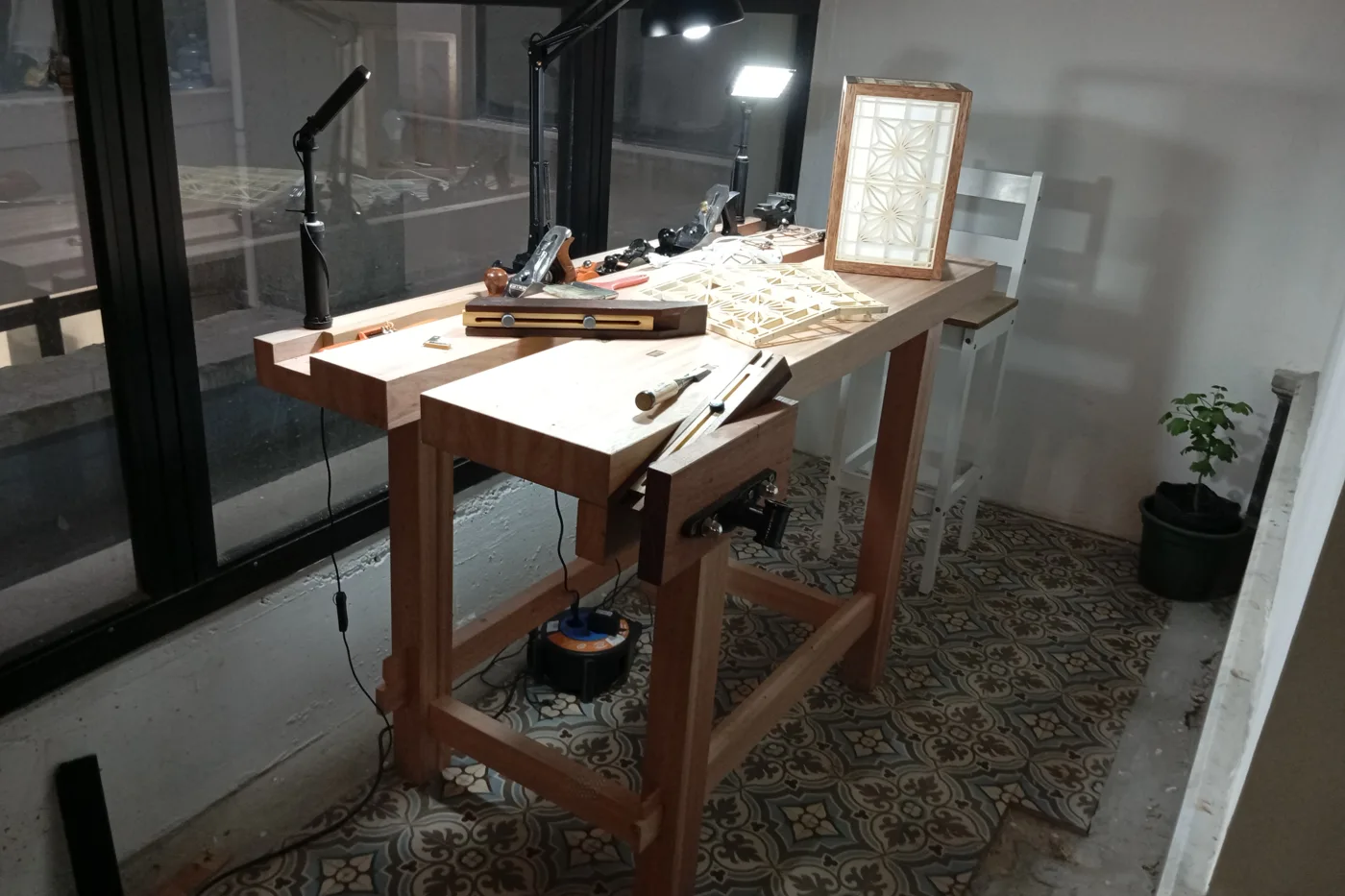 El banco de trabajo del taller, con cepillos, formones y una lámpara kumiko encendida sobre la mesa.